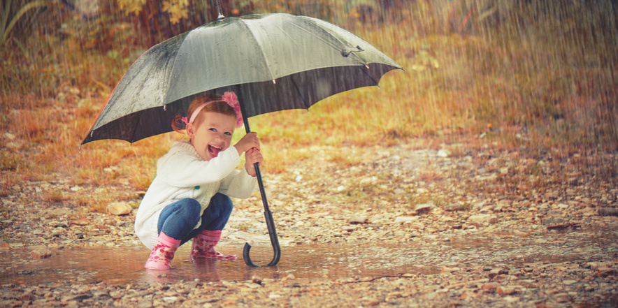 Kleines Mädchen kniet lachend und hält einen Regenschirm im strömenden Regen
