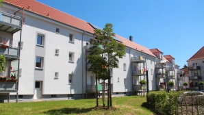 Hellgraues Wohnhaus mit neuem Dach, angebauten Balkone, 3 Etagen. Davor eine Wiese mit kleinen Bäumen. Hellgraues Wohnhaus mit neuem Dach, angebauten Balkone, 3 Etagen. Davor eine Wiese mit kleinen Bäumen.