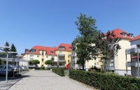 Mehrere sanierte Altbauten in gelb und hellbau, Blick auf einen gepflasterten Weg, ringsum viel Grün.