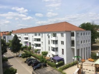 Wohnhäuser mit Satteldach, graue Fassade, weiße Balkonbrüstungen. Parkplätze mit Autos, rechts Spielplatz. 