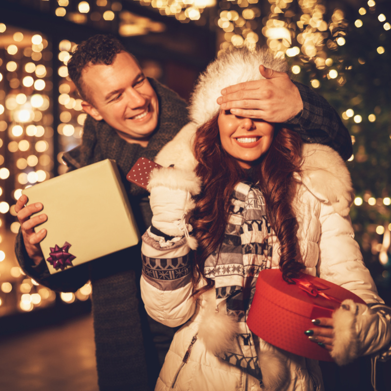 Ein Paar vor einem weihnachtlich beleuchtetem Schaufenster. Man hält Frau die Augen zu, um sie mit einem Geschenk zu überraschen.