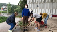 Workshop shore.oo Zwei Teilnehmer*innen führen einen großen Ring mit Blättern und Ästen bespannt an einer dritten Teilnehmerin auf und ab