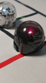 Die Szene zeigt zwei kleine, kugelförmige Roboter, die auf einem weißen Blatt positioniert sind. Der Roboter links ist transparent und der rechts ist tiefschwarz. Beide Roboter stehen auf einem weißen Hintergrund, auf dem schwarze Linien