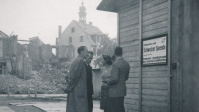 Fotoaufnahme der Baracke der Schweizer Hilfe auf dem Platz am Staatstheater vor der Trümmerlandschaft der Stadt. Im Hintergrund die Turmspitze der evangelischen Kirche.