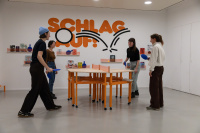 Schlag Auf! - Leo Scheidt Vier Personen, die Tischtennis spielen. Im Hintergrund eine weiße Wand mit dem orangen Schriftzug Schlag Auf! und Regale mit Büchern