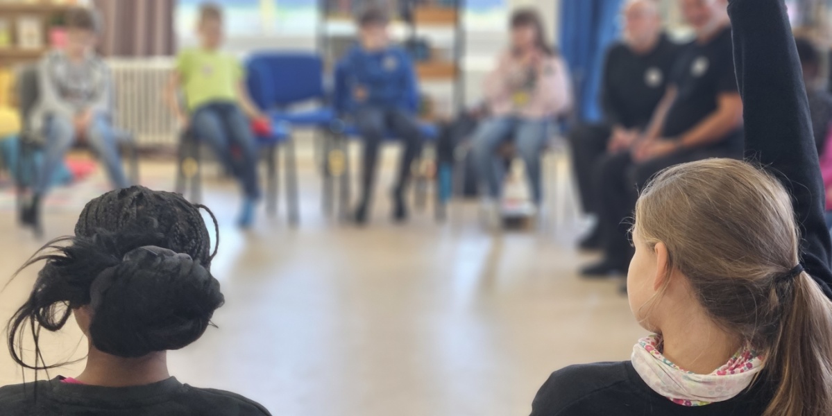 Kinder und Erwachsene sitzen im Stuhlkreis, ein Kind meldet sich