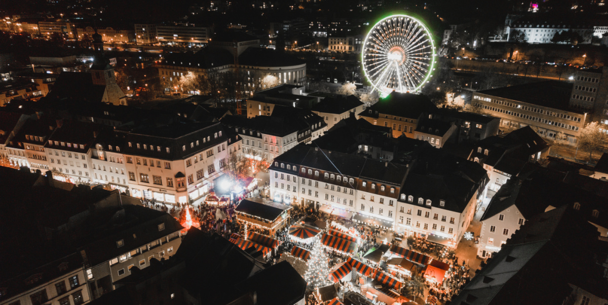 Luftaufnahme mit hell beleuchteten Buden und Riesenrad zwischen dunklen Hausdächern bei Nacht