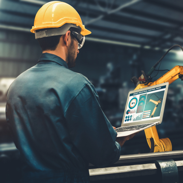 Mitarbeiter mit gelbem Helm und Laptop in einer Fertigungshalle mit gelben Roboterarmen