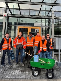 Sechs Personen mit orangefarbenen Warnwesten vor einem Gläsernen Gebäude, im Vordergrund ein Bollerwagen mit Reinigungsutensilien