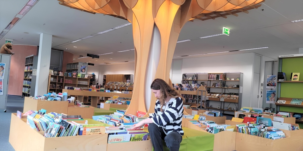 Baumstruktur in der Kinderbibliothek Eine Frau mit langen Haaren sitzt vor einem Bücherregal in einer Bibliothek, umgeben von bunten Kinderbüchern. Über ihr schwebt eine kunstvolle Baumstruktur.