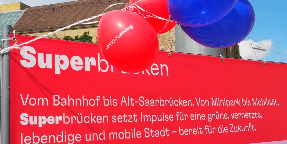 Rotes Banner mit rot-blauen Ballons - Aufschrift:  Vom Bahnhof bis Alt-Saarbrücken. Von Minipark bis Mobilität. Superbrücken setzt Impulse für eine grüne, vernetzte, lebendige und mobile Stadt – bereit für die Zukunft. 