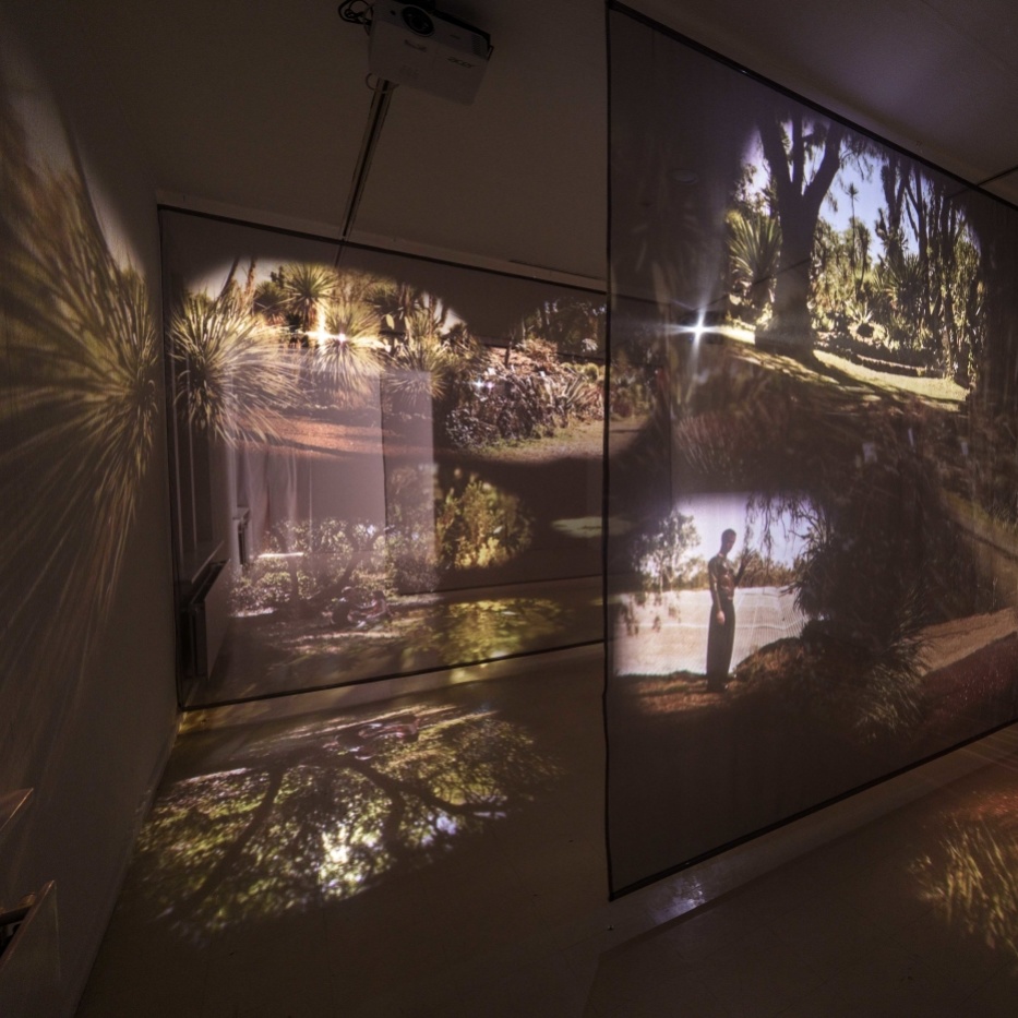 LOOPZONES Suzan Noesen Blick in einen abgedunkelten Ausstellungsraum in dem auf zwei transparenten Leinwänden ein Film zu sehen ist