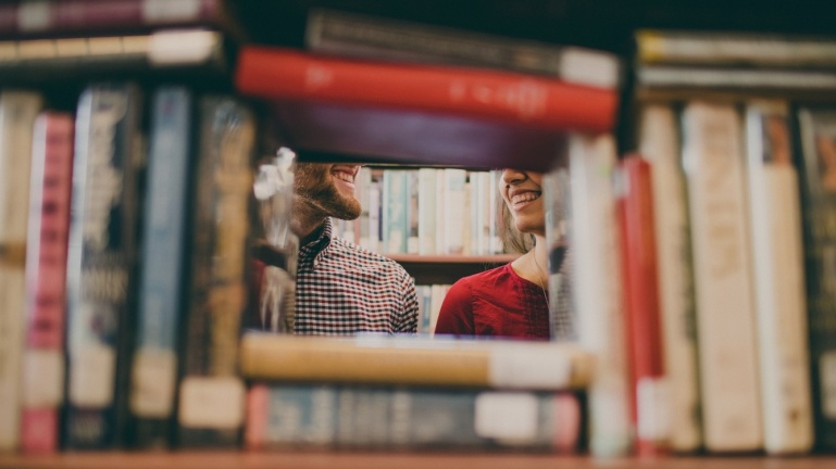 Zwei Personen lächeln sich durch eine Lücke zwischen den Büchern in einem Bibliotheksregal an.
