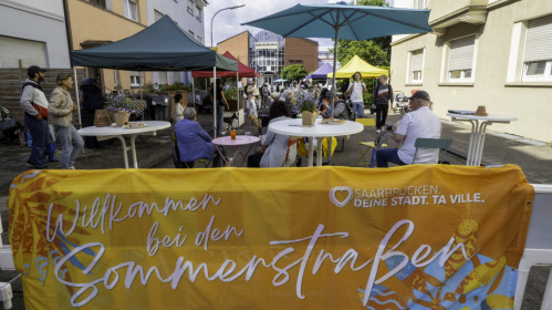 Eine Absperrung über die ein Banner mit der Aufschrift "Sommerstraßen" gespannt ist. Dahinter ist eine Straße mit vielen Passanten und bunten Schirmen zu sehen.