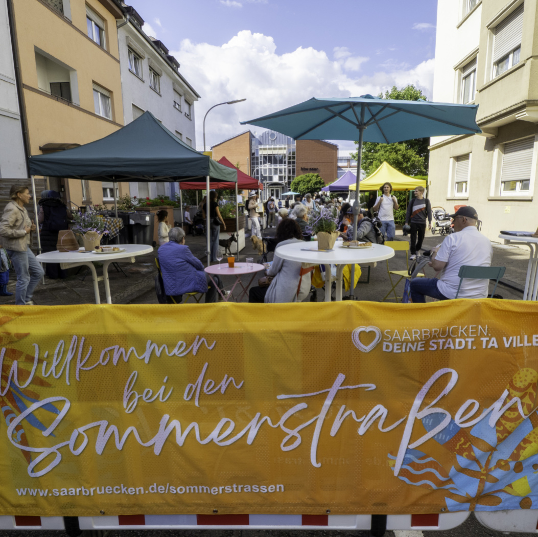 Vorne steht eine Absperrung über die ein Banner gespannt ist. Dahinter ist eine Straße mit vielen Passanten und bunten Schirmen zu sehen.