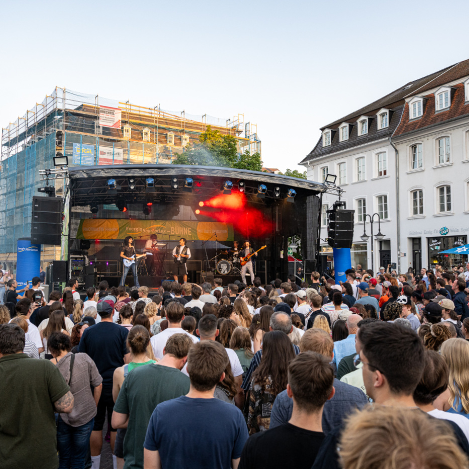Band auf einer Freiluftbühne vor zahlreichem Publikum, rote Bühnenbeleuchtung, Gebäude im Hintergrund.