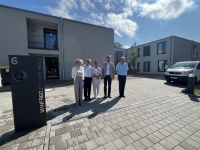 Eine Gruppe Menschen posiert neben einer Stele auf einem gepflasterten Hof vor einem Neubau 
