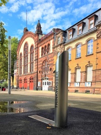 Im Vordergrund ist eine metallische Stele an deren Seite der Schriftzug Trinkbrunnen steht. Im Hintergrund sieht man ein altes großes Gebäude und Blattgrün.