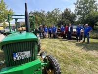 Hinter einem alten Traktor steht eine Gruppe Menschen auf einer Wiese