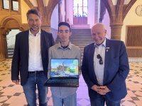 In einem alten Foyer stehen drei Männer. Der jüngste in der Mitte hält seinen geöffneten Laptop in die Kamera.