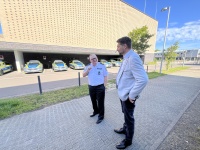 Zwei Männer stehen vor einem hellem, hallenartigen Gebäude unter dem viele Polizeifahrzeuge parken