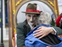 Geschminkter Schauspieler mit Hut umarmt ältere Frau herzlich