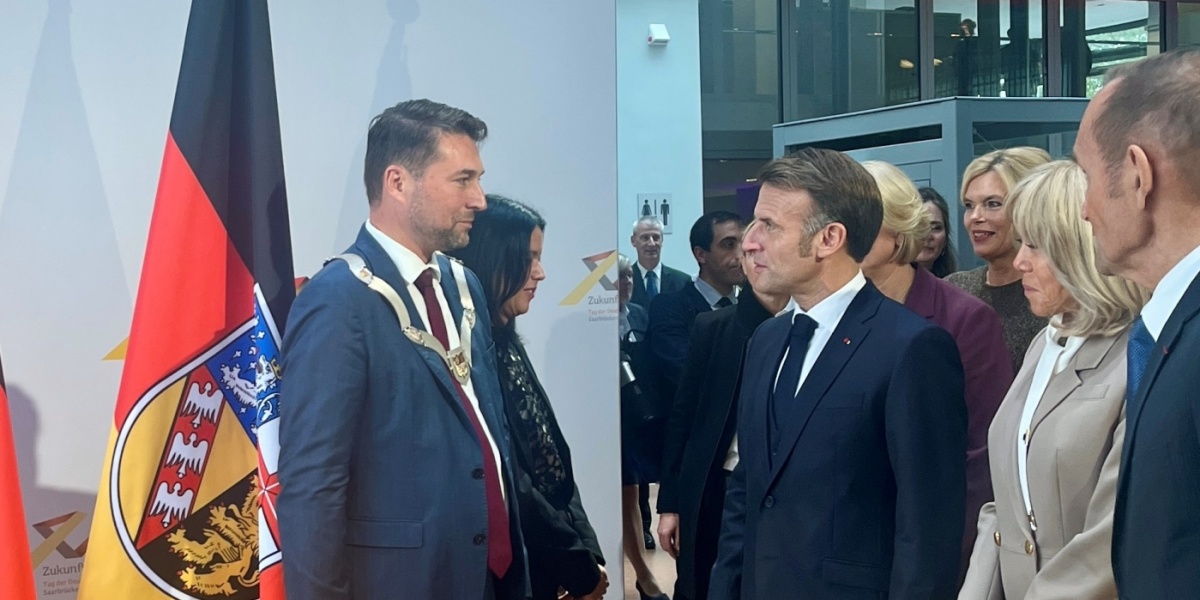 Eintrag ins Goldene Buch nach dem Festakt am Tag der Deutschen Einheit 2025 OB Uwe Conradt und der französische Präsident Emmanuel Macron stehen sich gegenüber, dahinter eine Saarland-Flagge