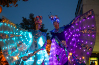 Nahaufnahme der beiden Stelzenläufer in farbig leuchtenden Kostümen mit Spiegelmosaik-Details und Kronen aus Lichtelementen, vor dunklem Abendhimmel.