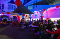 Menschen sitzen an Bierbänken in einem Innenhof, bunte Papierlaternen hängen über den Köpfen, die Wände sind in Blau und Rot angestrahlt.