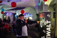 Ein Mann mit Basecap und weißem Bart spricht in ein Mikrofon und zeigt auf ein Banner mit der Aufschrift „0 % Gewalt“, Menschen sitzen und stehen im Hintergrund.