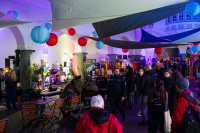 Blick über den bunt beleuchteten Innenhof mit vielen Menschen, einer Bühne mit Liveband und Verkaufsständen, bunte Laternen hängen unter den Stoffsegeln.