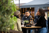 Drei Personen stehen an einem Hochtisch mit Gläsern, eine Pflanze im Vordergrund, im Hintergrund weitere Menschen.