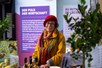 Eine Frau mit rotem Barett und gelbem Mantel steht hinter einem Stand mit Honig- und Weinprodukten, im Hintergrund Infotafeln.