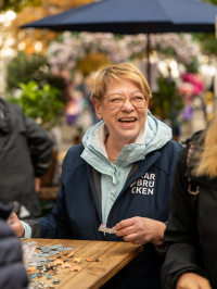Eine Frau mit Brille und blauer Jacke mit der Aufschrift „Saarbrücken“ steht an einem Holztisch mit Bastelmaterial und lacht.