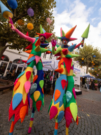 Zwei Stelzenläufer in bunten Kostümen mit spitzen Formen stehen vor einem Baum mit bunten Lampinons