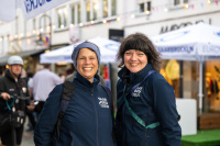 Zwei Frauen in blauen Jacken mit der Aufschrift „Saarbrücken“ stehen nebeneinander und lächeln in die Kamera, im Hintergrund weiße Sonnenschirme