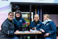 Vier Personen mit dunkel blauen Jacken mit der Aufschrift Saarbrücken stehen an einem tisch und lächeln in die Kamera