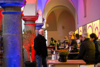 Mehrere Menschen sitzen und stehen an Tischen in einem Innenhof mit beleuchteten Arkaden und buntem Licht.