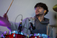 Ein Schlagzeuger mit Brille und Locken spielt an einem roten Schlagzeug, im Vordergrund Schlagzeugstöcke und Mikrofone.