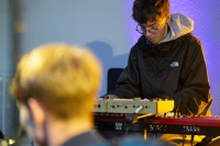 Ein Keyboardspieler mit Brille und schwarzer Jacke sitzt vor einem roten Keyboard, im Vordergrund unscharf ein Kopf mit blonden Haaren.