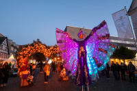 Bürgerfest zum Tag der Deutschen Einheit - Tag 3 am 4. Oktober auf der Festmeile der Eurometropole Ein Stelzenläufer in einem lila Kostüm mit leuchtenden Lichtern steht neben einem Blumenbogen aus Blüten, im Hintergrund Menschen.