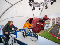 Kinder fahren auf Fahrradsimulatoren im Gesundheitszelt, im Hintergrund Modell eines großen roten Herzens der IKK Südwest.
