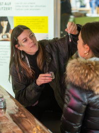 Künstlerin trägt Glitzerschminke bei einer Besucherin auf, Nahaufnahme am Stand der „Glitter Bar“.