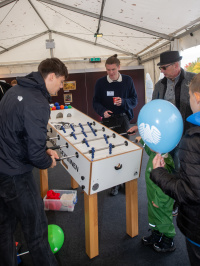 ugendliche und Erwachsene spielen gemeinsam Tischfußball an einem Kicker im Festzelt, Kinder halten Luftballons.