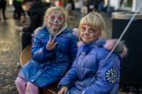 Zwei Kinder mit bemalten Gesichtern in blauen Winterjacken sitzen auf einer Bank, eines zeigt fröhlich das Peace-Zeichen.