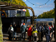 Stand der Bundesregierung mit Holzbogen-Eingang, Besucher stehen in der Sonne und warten am Dialogforum.