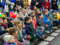 Gruppe von Kindern sitzt in der ersten Reihe und verfolgt gespannt eine Straßenaufführung, viele halten bunte Luftballons.