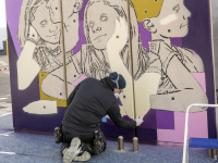 Künstler sprüht mit Spraydosen ein Wandbild mit Porträts von drei Kindern auf lila-gelbem Hintergrund.