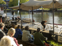 Künstler tritt barfuß auf einer schwimmenden Bühne an der Saar mit weißen Jonglierbällen auf, Publikum sitzt am Ufer.