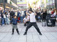 unge und Tänzer treten gemeinsam vor großem Publikum in der Innenstadt auf, Zuschauer beobachten die Szene.
